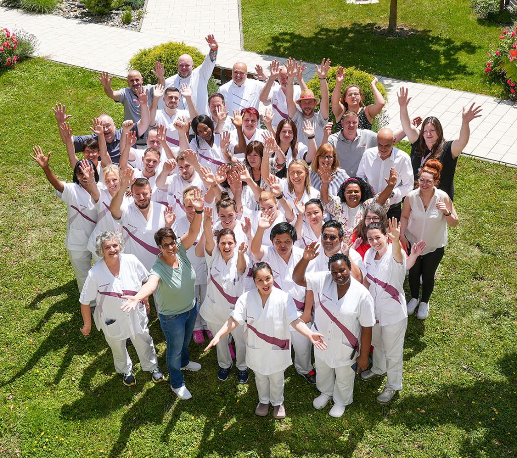 Das Foto zeigt eine Gruppe von Mitarbeiterinnen und Mitarbeitern des Maria Stadler Haus. Es wurde von einer erhöhten Position aufgenommen. Die Personen auf dem Foto winken in die Kamera.