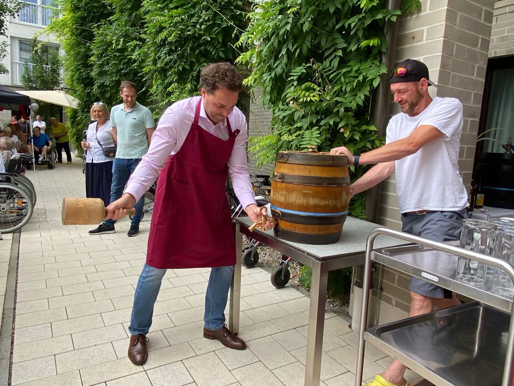 Das Foto zeigt zwei Männer bei einer Veranstaltung im Außenbereich des Maria Stadler Haus. Einer von ihnen ist dabei, mit einem Hammer ein Fass anzuzapfen.